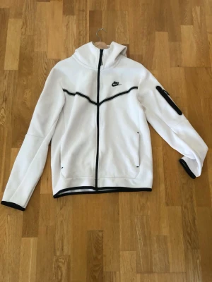 Vit Nike hoodie med svarta detaljer - Snygg vit hoodie från Nike med svarta detaljer och dragkedja framtill. Hoodien har huva, svarta linjer över bröstet och ärmslut samt en ficka med dragkedja på ena ärmen. Perfekt för en sportig och clean look.