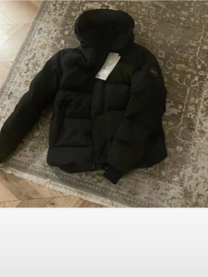 Svart dunjacka med huva Canada Goose - Säljer en svart dunjacka från Canada Goose med huva och diskret logotyp på ärmen. Jackan har en puffig design, dragkedja framtill och breda kanaler. Perfekt för kalla vinterdagar och riktigt snygg till streetwear-stilen. Är öppen till biliga pris då ja behöver pengar jackan är endast testad 1 gång och passar 170-180 cm de är bara att komma med bud men inga skambud 