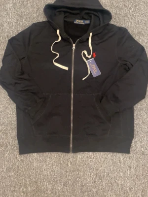 Svart hoodie från Polo Ralph Lauren - Snygg svart hoodie från Polo Ralph Lauren med dragkedja, vit dragsko och klassiska röda loggan broderad på bröstet. Hoodien har kängurufickor och är tillverkad i mjukt bomullsmaterial. Perfekt för en avslappnad och stilren look.