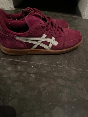 Vinröda Asics sneakers i mocka - Säljer ett par vinröda Asics sneakers i mocka med vita detaljer och klassisk logga på sidan. Skorna har bruna gummisulor och lågt skaft. Perfekta för dig som vill ha en snygg och bekväm sneaker med retrovibbar.