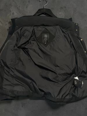 Svart dunjacka från Canada Goose - Säljer en svart dunjacka från Canada Goose med klassisk patch på ärmen. Jackan har en stilren design, hög krage och slät yta. Perfekt för kalla vinterdagar tack vare det varma dunfodret. Passar dig som vill ha både funktion och stil.