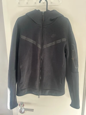 Svart Nike hoodie med dragkedja - Säljer en svart hoodie från Nike med hel dragkedja framtill och huva. Hoodien har diskret Nike-logga på bröstet, två fickor och en extra ficka med dragkedja på ärmen. Tillverkad i mjukt material, perfekt för en avslappnad och sportig stil.