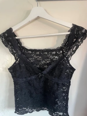 Svart spetslinnetopp med rosett - Säljer en svart, ärmlös linnetopp i spets med v-ringning och liten rosett framtill. Toppen har genomgående spetsdetaljer och en croppad passform som ger en trendig look. Perfekt att styla med höga jeans eller kjol för en cool outfit.