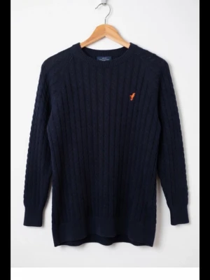 Mörkblå stickad tröja från Pier One storlek S - Säljer en mörkblå stickad tröja från Pier One med kabelstickat mönster och en liten röd broderad detalj på bröstet. Tröjan har långärmad design och ribbade muddar vid hals, ärmslut och nederkant. Perfekt för kyliga dagar.