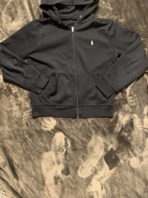 Svart hoodie från Polo Ralph Lauren - Snygg svart hoodie med dragkedja från Polo Ralph Lauren. Klassisk design med huva, fickor framtill och den ikoniska broderade loggan på bröstet. Tillverkad i mjukt bomullsmaterial som är skönt att bära. Perfekt för en avslappnad och stilren look.
