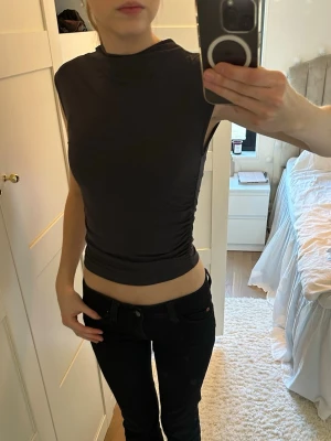 Svart tight ärmlös topp - Säljer en svart, tight och ärmlös topp med hög hals. Toppen har en slimmad passform och är gjord i ett stretchigt syntetmaterial som sitter snyggt mot kroppen. Perfekt att styla med jeans eller kjol för en clean look.