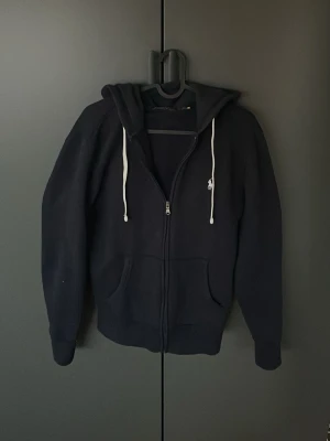 Mörkblå hoodie från Polo Ralph Lauren - Säljer en svart hoodie från Polo Ralph Lauren med dragkedja, vit broderad logga på bröstet och vita dragsnören i huvan. Hoodien har fickor framtill och är gjord i mjukt bomullsmaterial. Perfekt för en chill och stilren look.