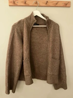 Beige kofta från Gina Tricot - Mysig beige kofta från Gina Tricot med öppen front, ribbad krage och två stora fickor framtill. Koftan är stickad i mjukt material och har långa ärmar samt ribbade muddar vid ärmslut och nederkant. Perfekt för lager på lager och chill stil.