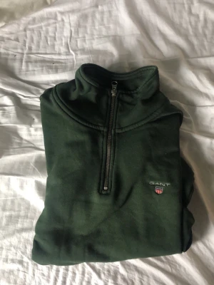 Mörkgrön half-zip tröja från Gant - Mörkgrön sweatshirt från Gant med halv dragkedja och ståkrage. Tröjan har broderad logga på bröstet och ribbade muddar vid ärmslut och nederkant. Perfekt för en avslappnad men snygg stil.