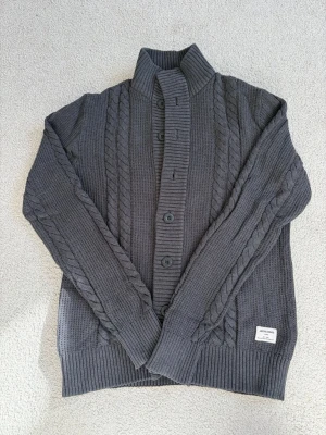 Mörkgrå kabelstickad kofta Jack & Jones - Mörkgrå kofta/cardigan från Jack & Jones.                   Storlek: S.                                                                               Färg: mörkgrå.                                                                     Skriv vid frågor, priset går även att diskuteras