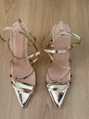 Guldiga pumps med remmar och klack - Säljer ett par glansiga guldiga pumps med spetsig tå och flera tunna remmar som går runt foten och ankeln. Skorna har hög klack och öppen häl, vilket ger en elegant och trendig look. Perfekta för att sticka ut och ge outfiten extra glam.