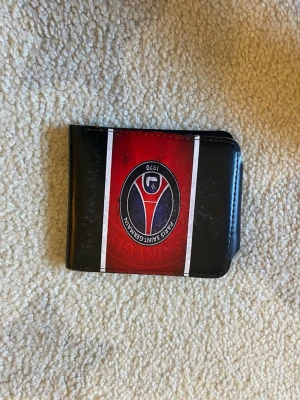PSG plånbok i svart och rött - Snygg plånbok från Paris Saint-Germain i svart och rött konstläder. Framsidan har PSG:s logga och vita ränder. Insidan har flera kortfack och ett fönster för ID eller busskort. Perfekt för dig som älskar fotboll och vill visa ditt lag.