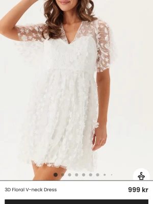 Vit kort blommig klänning med spets - Söt vit kort klänning med v-ringning och transparenta ärmar dekorerade med blommig spets. Klänningen har 3D-blommor över hela tyget och en lätt utsvängd kjol. Perfekt för dig som vill ha en romantisk och trendig look. PERFEKT STUDENTKLÄNNING. Aldrig använd utan endast testad. Säljer för att jag tyvärr valde en annan. 