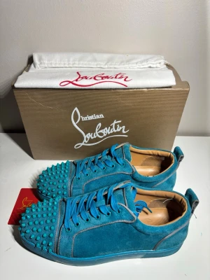 Blåa spikes-sneakers från Louboutin - Unika blå sneakers från Christian Louboutin i mocka med matchande blå skosnören och coola spikes på tån. Låg modell med exklusiv känsla och läderfoder. Perfekta för dig som vill sticka ut med stilrena detaljer och lyxig design. Dustbag och box och annat tillbehör medföljer, tror inte dom är äkta