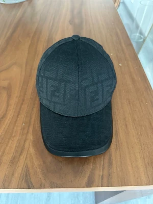 Svart keps från Fendi med logomönster - Säljer en svart keps från Fendi med diskret logomönster över hela kepsen. Justerbar rem baktill med metallspänne och Fendi-tryck. kepsen har klassisk rundad form och böjd skärm. Perfekt för dig som gillar exklusiva accessoarer.
