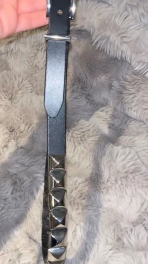 Svart bälte med silverfärgade nitar - Coolt svart bälte med stora pyramidformade silvernitar längs hela bandet. Bältet har ett klassiskt spänne i metall och är tillverkat i ett läderliknande material. Perfekt för att ge din outfit en edgy touch.