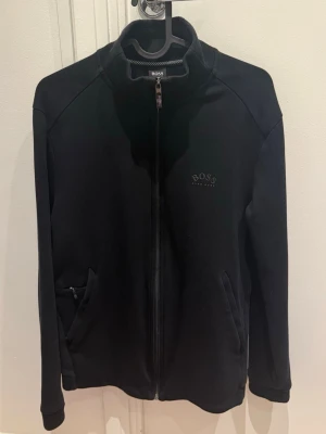 Svart zip-hoodie från BOSS - Stilren svart hoodie från BOSS med dragkedja framtill och diskret logga på bröstet. Tröjan har två fickor med dragkedja och en klassisk, sportig look. Perfekt för en avslappnad och trendig stil.