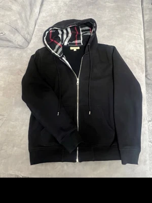 Svart Burberry zip hoodie - Säljer en svart hoodie med dragkedja från Burberry. Tröjan har klassiskt rutigt foder i huvan och längs insidan, samt två fickor framtill och justerbar dragsko i huvan. Perfekt för dig som gillar streetwear och vill ha något stilrent med en twist.