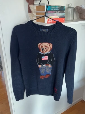 Mörkblå stickad tröja Polo Bear - Mörkblå stickad tröja från Polo Ralph Lauren med ikoniskt Polo Bear-motiv på bröstet. Tröjan har rund hals, långa ärmar och ribbade muddar. Använd endast en gång och skriv gärna vid funderingar 