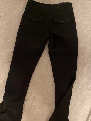Gina tricot- kostymbyxor-dam - Svarta kostymbyxor  med slim fit och två bakfickor med lock och knapp. Kostymbyxorna har normal midja och flared ben.  Materialet ser stretchigt ut vilket ger en skön passform. Perfekta för en clean och modern stil. Lite utslitna vid fötterna . 