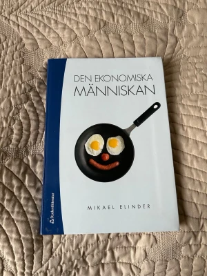 Den ekonomiska människan - Kurslitteratur i nationalekonomi/företagsekonomi skriven av Mikael Elinder. Boken har ett vitt och blått omslag med en stekpanna och mat som motiv. Perfekt för studenter som vill förstå grundläggande ekonomiska teorier och mänskligt beteende.