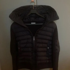 Svart Moncler pufferjacka med huva - Svart pufferjacka från Moncler med stickade ärmar och quiltad kropp. Jackan har en stor huva, dragkedja framtill och praktiska fickor med knappar och dragkedja på ärmen. Perfekt för dig som vill ha en snygg och sportig look med exklusiv känsla.