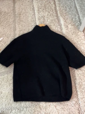 Svart stickad tröja med halvpolokrage - En svart stickad tröja med halvpolokrage och korta ärmar. Tröjan har en rak passform och är tillverkad i ett mjukt material som passar perfekt för lager på lager. Enkel och stilren design som funkar till det mesta.