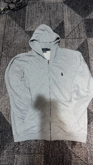 Grå hoodie från Polo Ralph Lauren - Snygg ljusgrå hoodie med dragkedja från Polo Ralph Lauren. Klassisk design med huva, fickor framtill och den ikoniska mörkblå loggan broderad på bröstet. Tillverkad i mjukt bomullsmaterial som känns skönt mot huden. Perfekt för en avslappnad och stilren look.