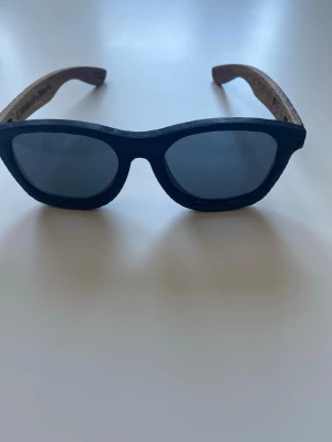  solglasögon i trä  - Snygga solglasögon från Woodzee med svarta glas och bågar i trä. Klassisk form med en cool touch av naturligt trä på skalmarna. Perfekta för dig som vill sticka ut med en unik accessoar.