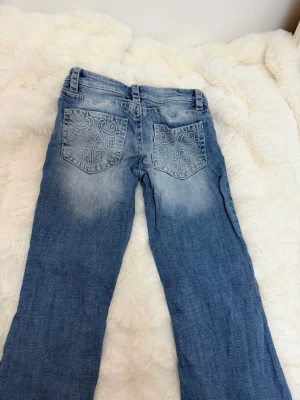 Blå bootcut jeans med broderade fickor - Blå bootcut jeans i denim med snygg ljus tvätt och broderade detaljer på bakfickorna. Klassisk femficksmodell med knapp och dragkedja framtill. Jeansen har en avslappnad passform och är perfekta för en trendig och ungdomlig stil.