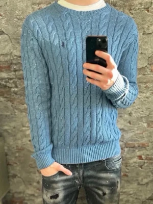 Blå kabelstickad tröja Polo Ralph Lauren - Kabelstickad blå tröja från Polo Ralph Lauren med rund halsringning och diskret broderad logga på bröstet. Tröjan har ribbade muddar och nederkant, perfekt för lager-på-lager-stil.