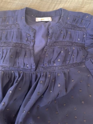 Mörkblå blus med volangdetaljer - Snygg mörkblå blus med volangdetaljer på axlarna och små broderade prickar över hela tyget. Blusen har rund halsringning och knappar framtill. Tillverkad i ett tunt, luftigt material som ger ett flowy intryck.