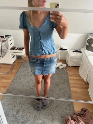 Blå topp med puffärm och knappar - Söt blå topp i denimliknande material med puffiga korta ärmar och knappar framtill. Djup v-ringning och lätt rynkad nederkant ger en avslappnad och trendig look. Perfekt till jeans eller kjol för en cool vardagsstil.