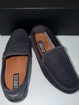 Mörkblå loafers från Limited - Snygga mörkblå loafers från Limited i mockaimitation med kontrastsömmar och perforerad ovandel. Skorna har en klassisk slip-on design och innersula i brunt konstläder för extra komfort. Perfekta för en stilren och avslappnad look.