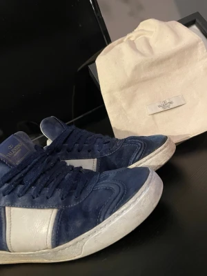 Blåa Valentino sneakers i mocka - Snygga blåa sneakers från Valentino med vita detaljer på sidan och vit sula. Skorna är låga och har en ovandel i mocka och läder. Klassisk design med Valentino-logga på plösen och matchande blå skosnören. Perfekta för dig som gillar exklusiva sneakers.