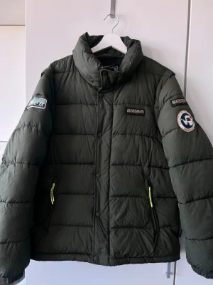 Mörkgrön pufferjacka och väst från Napapijri - Säljer en mörkgrön pufferjacka från Napapijri med hög krage och broderade loggor på bröstet och ärmen. Vinterjackan kan man också ha som väst! Luvan går också att ta bort om önskas. 