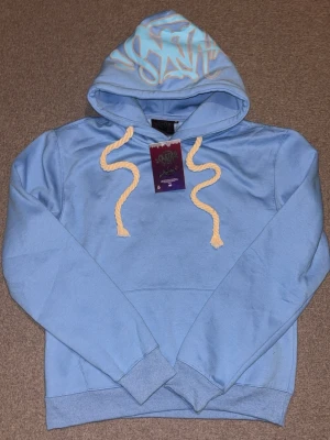 Ljusblå hoodie från syna - Snygg ljusblå hoodie från Syna med stor huva och graffiti-inspirerat tryck i vitt och beige på huvan. Hoodien har grova, tvinnade snören i beige och ribbade muddar vid ärmar och nederkant. Perfekt för en avslappnad streetwear-look.