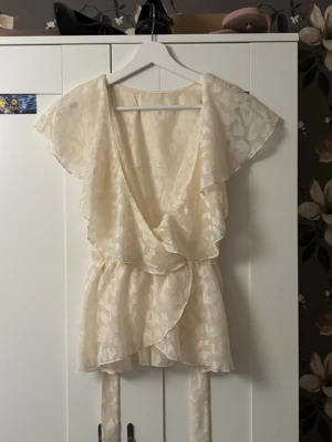 Krämvit omlottblus med volanger - Söt krämvit omlottblus med volangdetaljer och knytband i midjan. Blusen har korta, fladdriga ärmar och ett lätt transparent tyg med diskret mönster. Perfekt att styla med jeans eller kjol för en trendig look. Den är jättefin men tycker inte om hur den sitter på mig. Den har typ nåt märke på insidan av tröjan där fram men jag har aldrig använt den men antar att det hänt i produktionen. Det syns dock inte när man har på sig den.