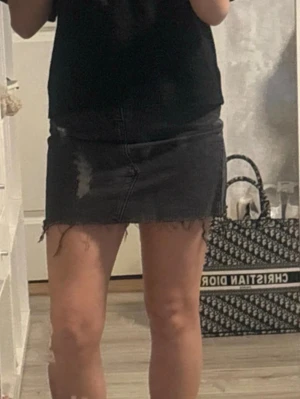 Svart jeanskjol med rå kant - Säljer en svart jeanskjol i minilängd med rå, fransig kant och slitningar framtill. Kjolen har klassisk femficksmodell, bälteshällor och knappgylf. Perfekt till en avslappnad och trendig look.