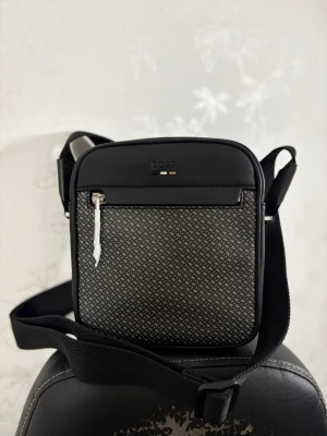 BOSS Hugo Boss crossbody väska – helt ny – premium design - Säljer en äkta BOSS Hugo Boss crossbody-väska.  Väskan är helt ny och oanvänd. Den köptes för bara några dagar sedan men visade sig tyvärr inte vara riktigt min stil.  Kan skickas direkt. Öppen för seriösa bud.  Nypris (typiskt)  ca 1 300 – 2 200 kr   Prislappen finns inte kvar eftersom jag tog bort den när jag först trodde att jag skulle börja använda väskan. Den har dock aldrig använts och är i nyskick.  Detaljer:  • Original BOSS-design • Stilren monogramfront • Justerbar axelrem • Flera fack