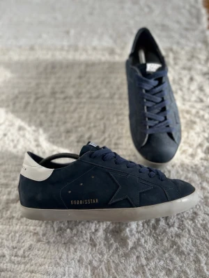 Mörkblå GGDB sneakers med vit detalj - Snygga mörkblå sneakers från Golden Goose med vit hälflik och GGDB/SSTAR-detalj i guld på sidan. Skorna har klassisk låg profil, stjärndesign på sidan och är gjorda i mocka med matchande mörkblå snören och vit sula. Storlek 43 true to size. Pris ej hugget i sten.