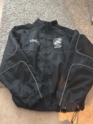 Svart vindjacka USA91 Sport - Snygg svart vindjacka från USA91 Sport med vita detaljer och dragkedja framtill. Jackan har hög krage, elastiska muddar och tunnt, lätt material med diskret mönster. Perfekt för aktiva dagar och streetwear-stil.