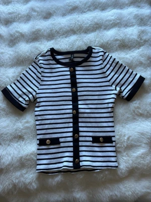 Randig kortärmad topp med knappar - Säljer en stilren randig topp med korta ärmar och dekorativa guldknappar framtill. Toppen har svarta och vita ränder, svarta detaljer vid ärmslut och halsringning samt två fejkfickor framtill. Perfekt för en trendig och klassisk look.