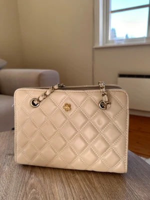 Beige quiltad handväska med kedja - Stilren beige handväska med quiltat mönster och guldfärgade kedjedetaljer. Väskan har en rektangulär form och är tillverkad i glansigt material som liknar skinn. Perfekt för dig som gillar klassisk och elegant stil.