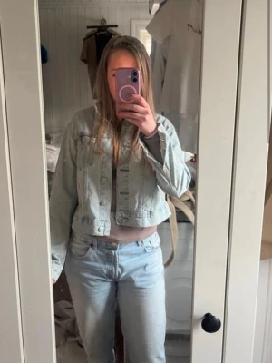 Ljusblå cropped jeansjacka - Trendig ljusblå jeansjacka i cropped modell med klassisk krage och knappstängning framtill. Jackan har två bröstfickor med lock och knappar samt långa ärmar. Perfekt för en avslappnad och cool stil.