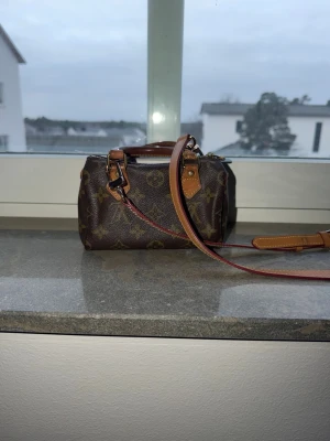 Louis Vuitton monogram handväska - Säljer en brun handväska från Louis Vuitton med klassiskt monogrammönster och detaljer i ljusbrunt läder. Väskan har både handtag och en avtagbar axelrem i läder, samt guldfärgade metalldetaljer. Perfekt storlek för det viktigaste och en ikonisk accessoar.
