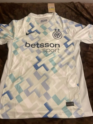 Inter Milan bortatröja Nike - Säljer en Inter Milan bortatröja från Nike med geometriskt mönster i vitt, ljusblått och blått. Tröjan har klubbmärke och Nike-logga på bröstet samt sponsortryck 'betsson sport' framtill. Tillverkad i lätt och ventilerande material, perfekt för fotboll.