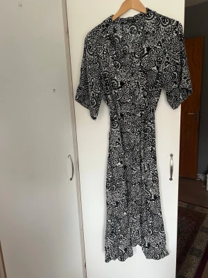 Svartvit mönstrad klänning  - Svartvit klänning med blommigt mönster och korta ärmar. Klänningen har v-ringning och knyt i midjan för en smickrande passform. Perfekt för dig som gillar en feminin och trendig stil.