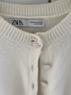 Vit kofta med pärlknappar från Zara - Supersöt vit kofta från Zara med rund halsringning och dekorativa pärlknappar framtill. Koftan är stickad i ett mjukt material och har långa ärmar samt ribbade muddar vid hals, ärmslut och nederkant. Perfekt för en chic och stilren look.