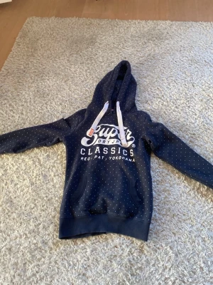 Blå Superdry hoodie med prickar XS - Mörkblå hoodie från Superdry med vita småprickar och stor vit logga på bröstet. Klassisk känguruficka framtill och vita dragsnören i huvan. Mjuk insida och ribbade muddar vid ärmar och nederkant. Perfekt för en avslappnad streetstyle.